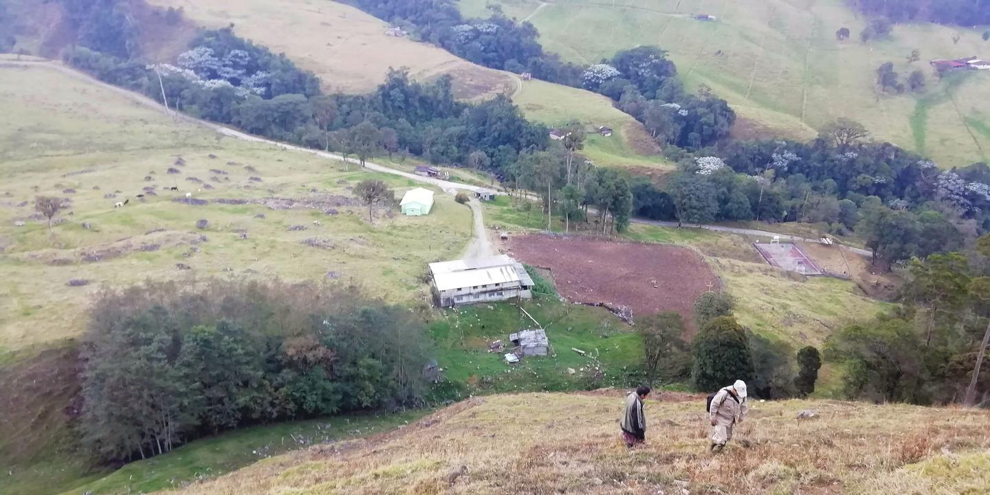 Vista desde una loma hacia una finca con techo de zinc; dos personas caminan por el pasto.