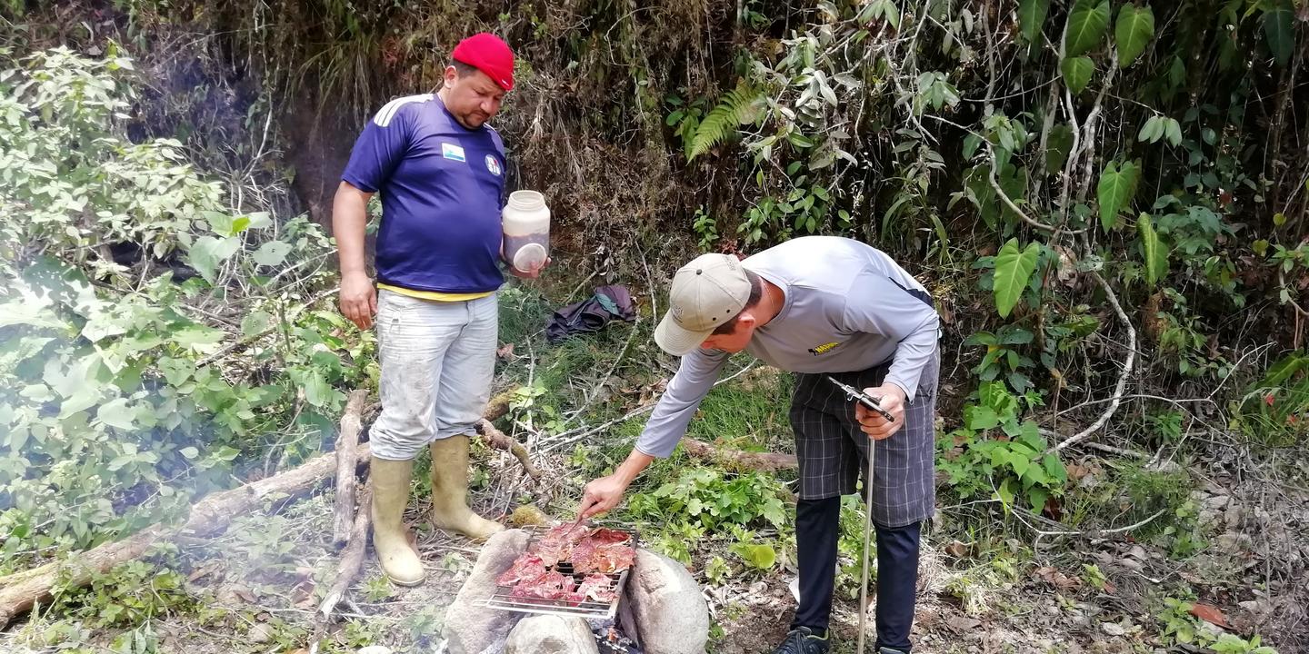 Dos hombres preparan carne asada en un fogón a la orilla del camino.