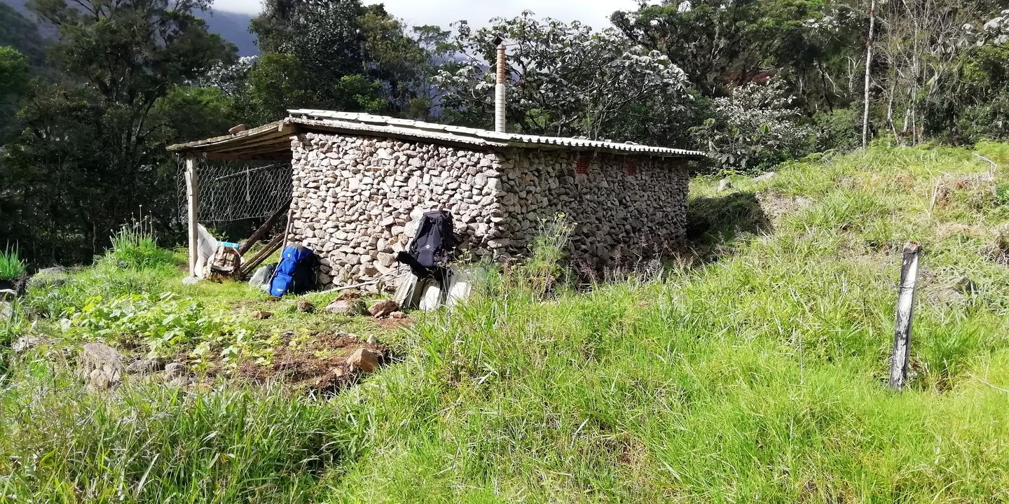 Una casita de piedra con techo de láminas; los morrales recostados a la pared.