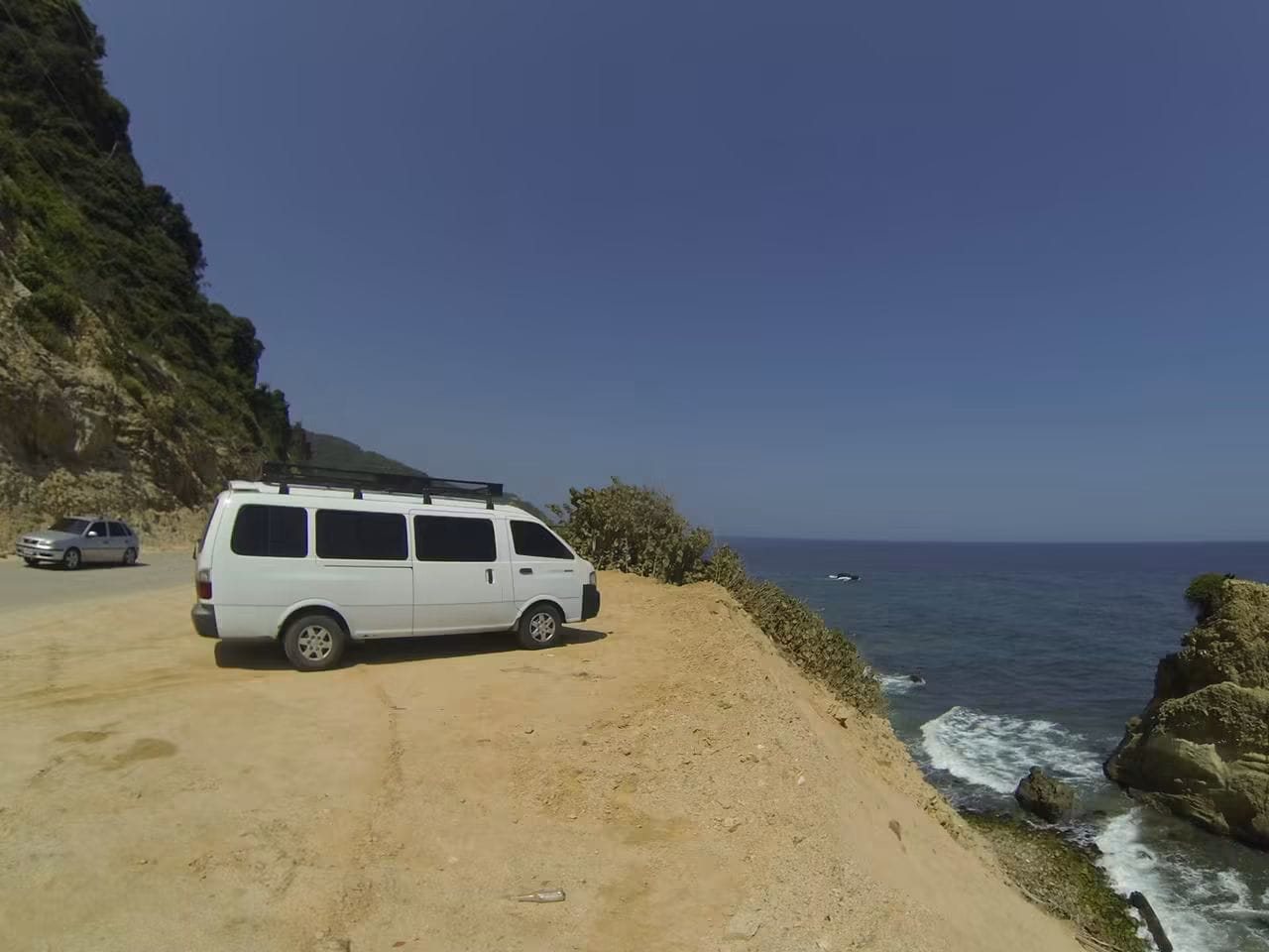 La Kia Pregio blanca, con parrilla en el techo, estacionada al borde de un acantilado frente al mar.